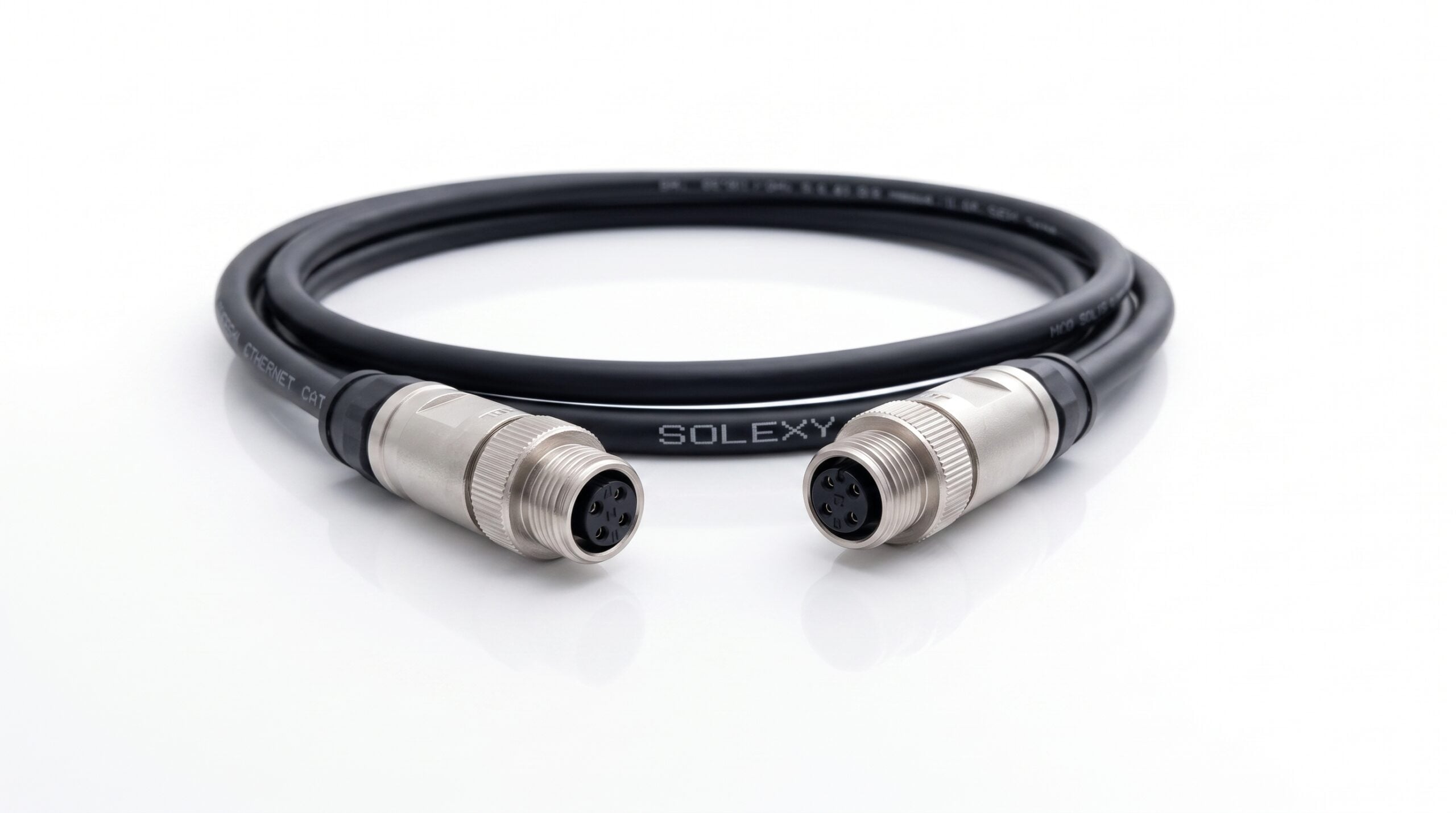 A coiled black industrial Ethernet cable featuring "SOLEXY" and "INDUSTRIAL ETHERNET CAT." text is centered against a reflective white background. The cable terminates in two prominent, threaded silver M12 connectors, with their 5-pin inserts and central keying prominently facing forward in sharp focus. The reflection of the connectors and coiled cable is visible on the glossy surface.